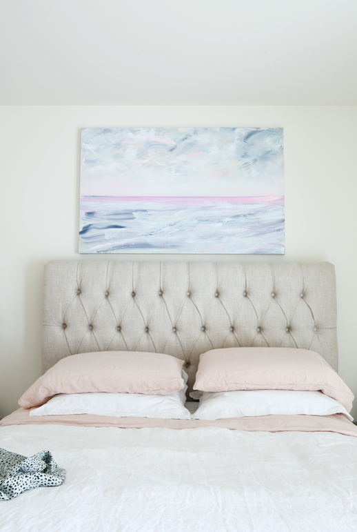 Photo shows a soft ocean landscape painting installed above a tufted queen sized headboard.  Blush pink and white sheets compliment the painting, which is soft and whimsical.  