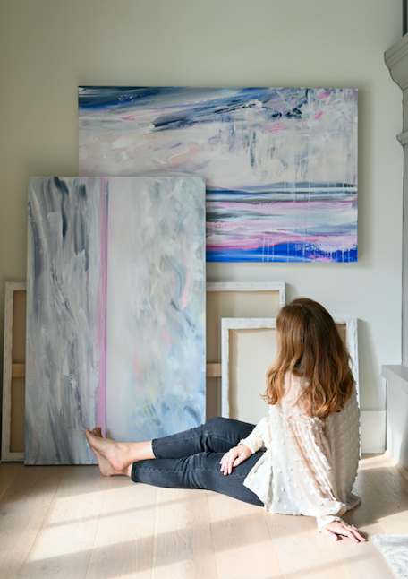 Artist Dana Mooney sits in front of her new collection of ocean landscape paintings, with her back to camera looking at the artwork.  Dana wears dark grey denim skinny jeans, bare feet and a sheer textured beige dotted flowy shirt.  