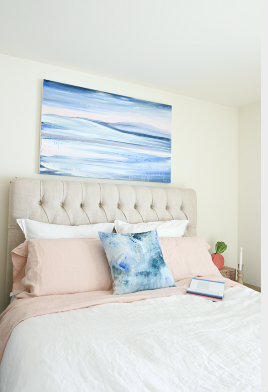 An abstract ocean landscape painting is hung on the wall above a tufted headboard.  White and blush pink bedding make this bedroom very soft and inviting.  