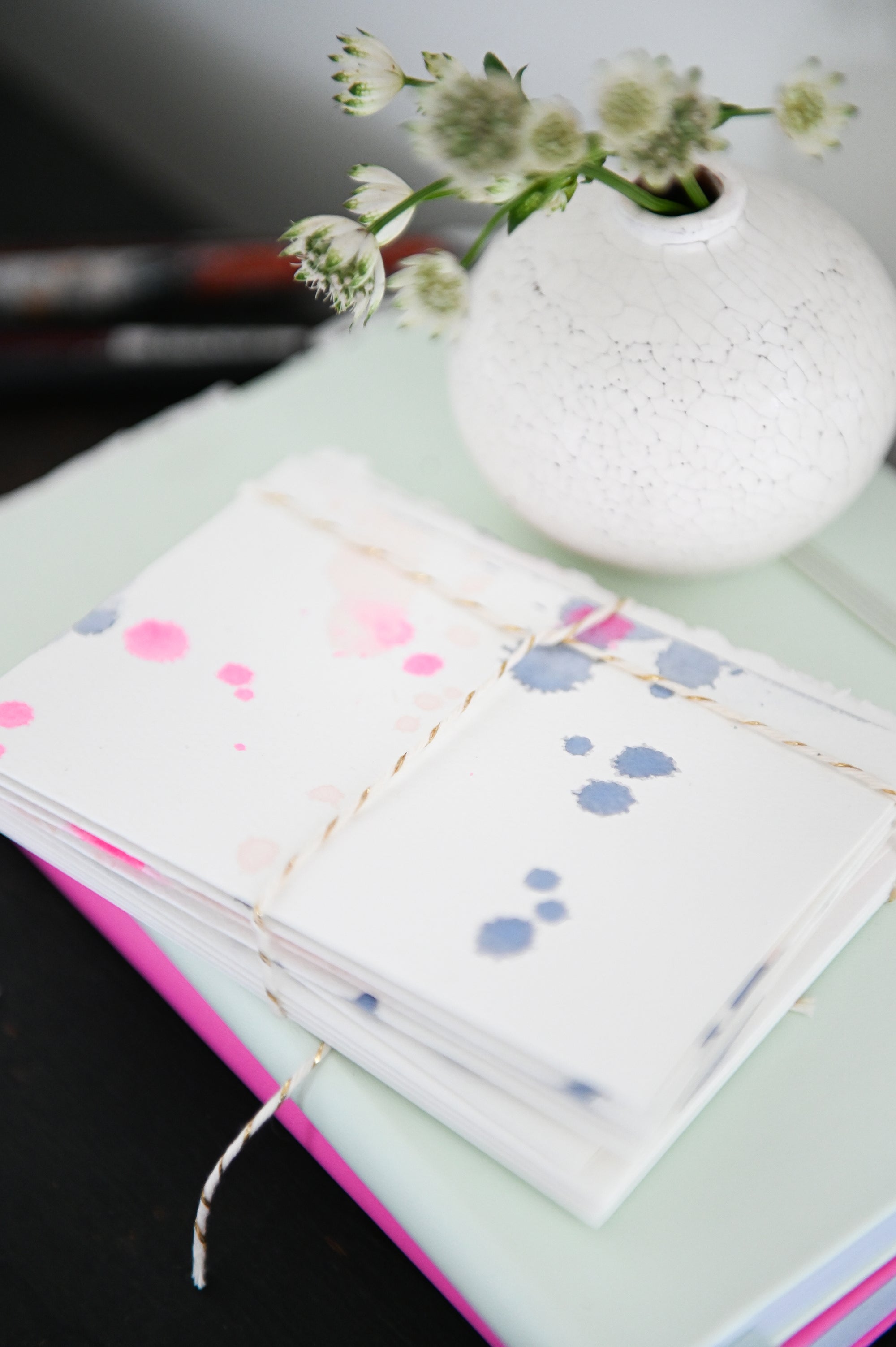 A stack of hand painted blank note cards sitting on top of a mint green and pink notebook, next to a small ceramic bud vase.  The cards are splatter paintied minimally with pink and blue watercolour dots.