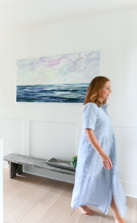 Artist Dana Mooney walks past her Installed Ocean Landscape painting, wearing a pale checkered oversized shirt dress.  This photo shows an ease and calmness that Dana's artwork ensues. 