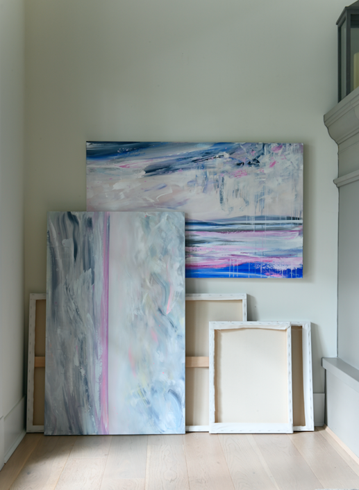A styled artist studio scene showcasing a collection of paintings on canvas, stacked randomly agains a wall.  One painting hangs horizontally while another is stacked vertically against it.  Some canvases are showing their front, some you can only see the raw canvas back.  Beside the canvases is a large cement fireplace mantle, and light wood flooring is lit by a window from the side. 