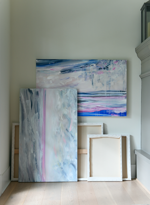 A styled artist studio scene showcasing a collection of paintings on canvas, stacked randomly agains a wall.  One painting hangs horizontally while another is stacked vertically against it.  Some canvases are showing their front, some you can only see the raw canvas back.  Beside the canvases is a large cement fireplace mantle, and light wood flooring is lit by a window from the side. 