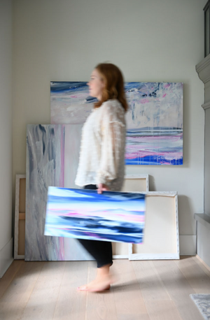 Artist Dana Mooney is blurred in the foreground as she walks past a stack of her canvas paintings leaning against the wall behind her.  She is holding a small landscape painting in her hand facing the camera, with pops of bright blues and pinks.