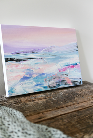 A small modern pastel ocean landscape painting sits upon a distressed wooden table.