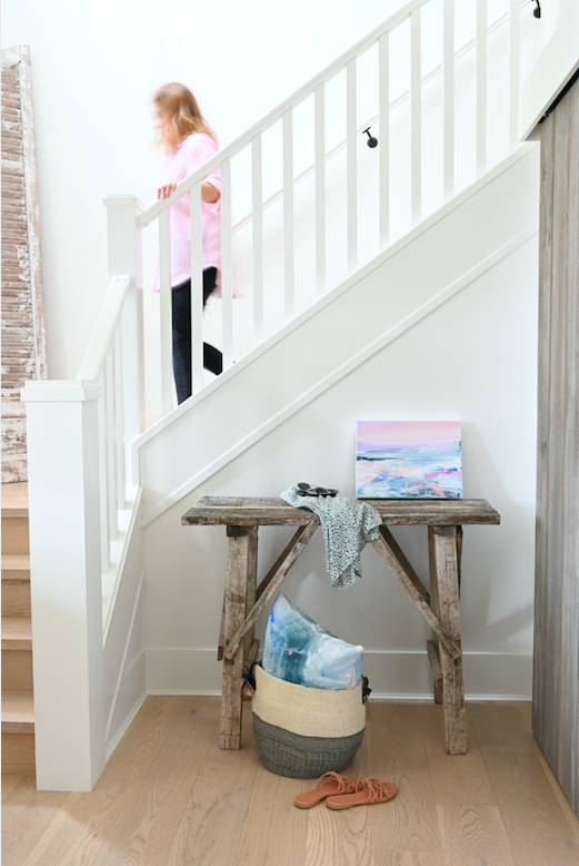 Artist Dana Mooney descends a staircase above a console table which holds one of her small landscape paintings.  The home is white and clean, with light wood floors and the painting is the pop of colour in the room.  Accent pieces of a basket and sandals upon the floor help to style the space and invite the viewer to imagine living with Dana Mooney's artwork.